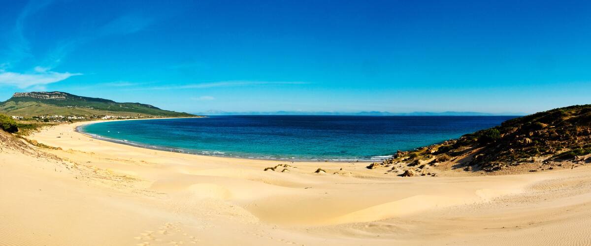 A coastal landscape with a sandy beach, a clear blue sea, and a hill with sparse vegetation.