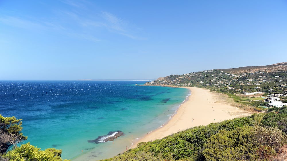 Eine Küstenlandschaft mit Sandstrand, klarem türkisblauem Wasser und einer hügeligen Kulisse mit vereinzelten Gebäuden.