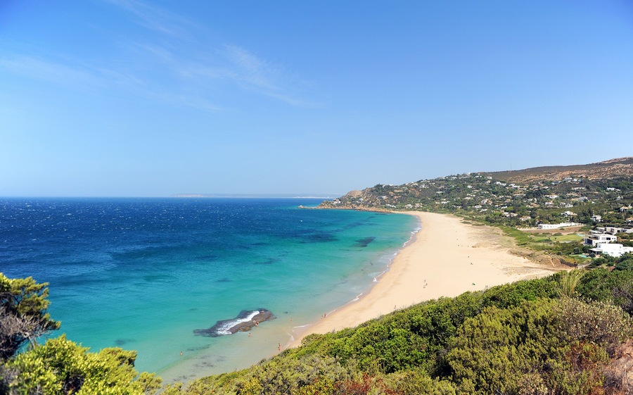 Paisaje costero con playa arenosa, aguas turquesas cristalinas y un fondo montañoso con edificios dispersos.