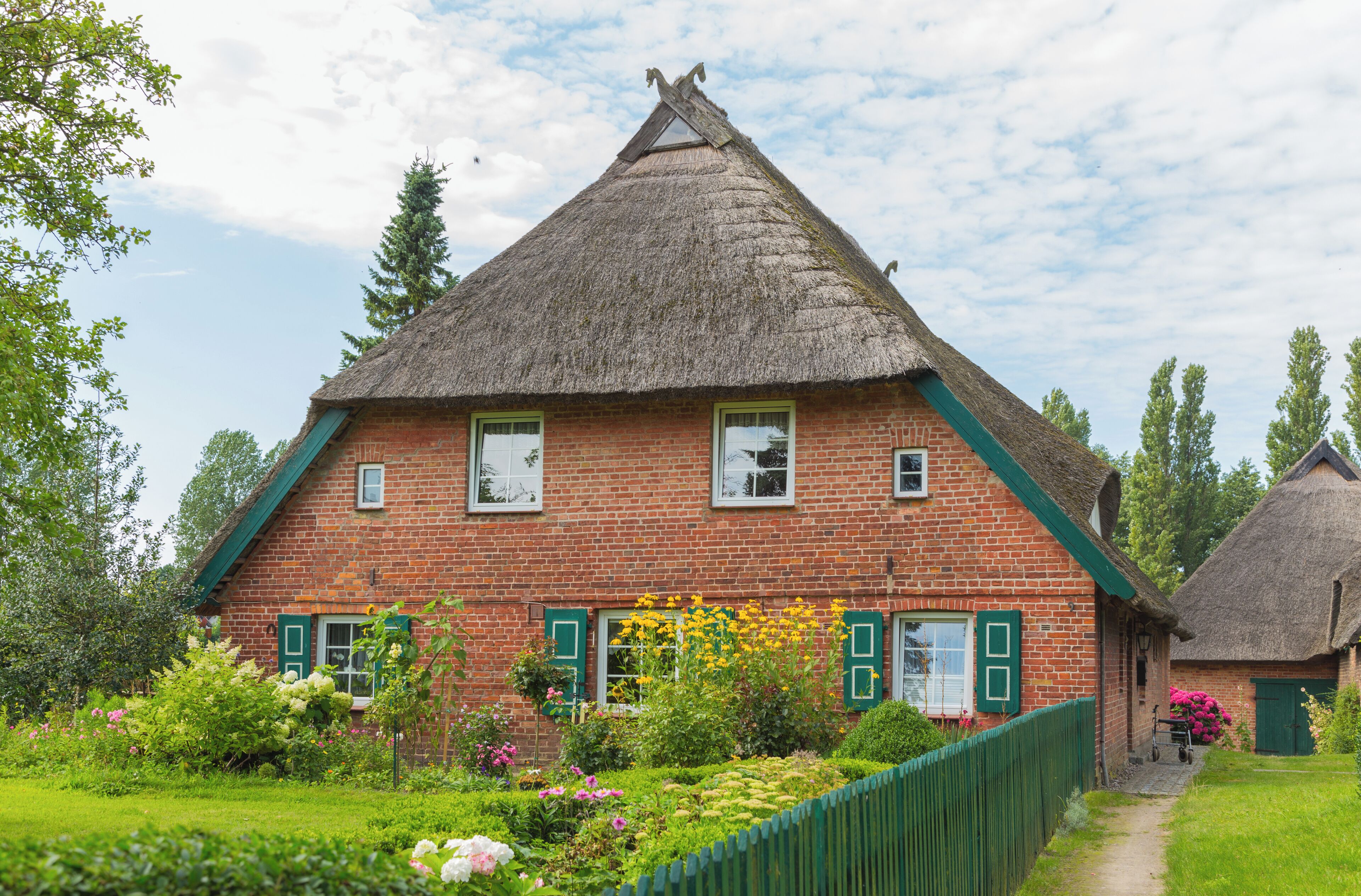 A traditional brick house with a thatched roof, surrounded by a lush garden and a wooden fence.