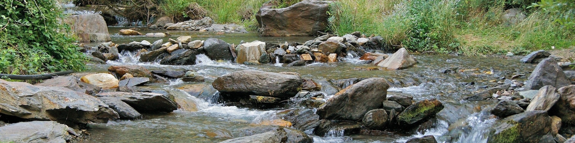 Un arroyo pedregoso con agua corriente, rodeado de vegetación y grandes rocas.