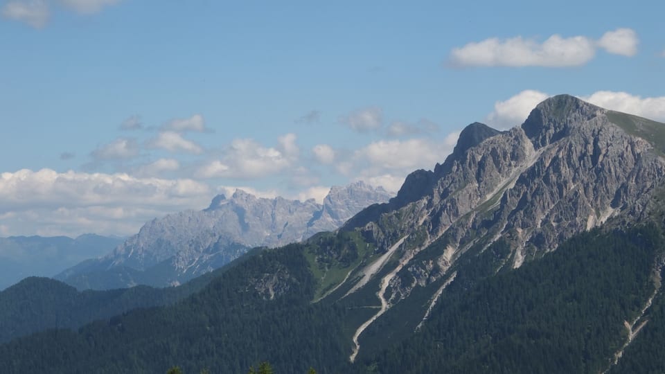 A mountain range with a prominent peak, dense forested slopes, and distant mountain ranges under a clear sky.