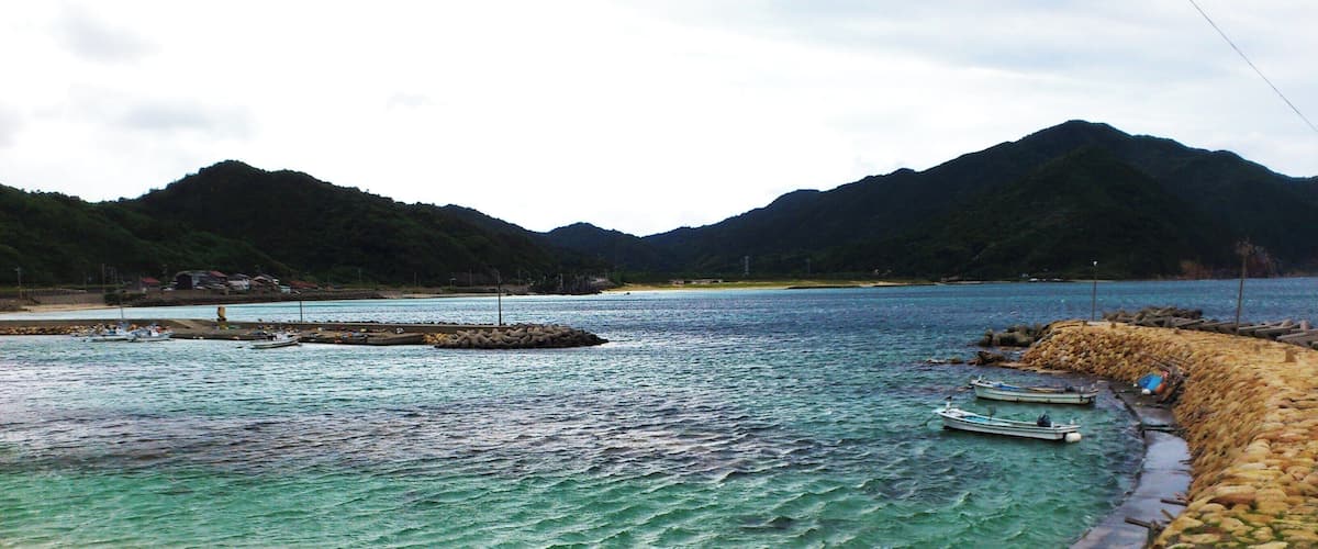 A coastal landscape with clear turquoise waters, a stone jetty, boats, and a mountainous backdrop.