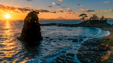 Um pôr do sol costeiro com uma formação rochosa em primeiro plano e uma montanha ao fundo.