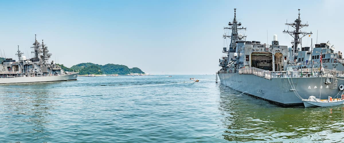 A naval ship docked at a port with another ship in the background.