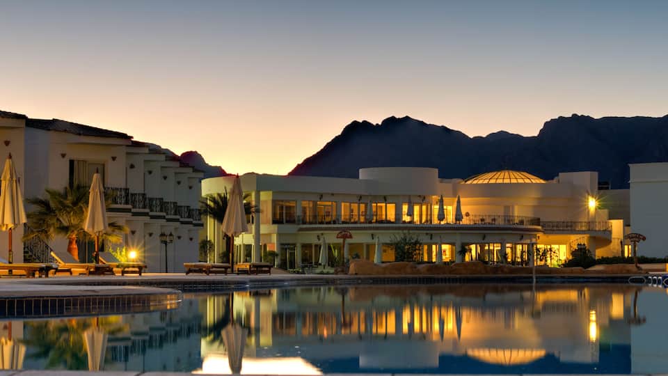 A resort with a swimming pool, lounge chairs, umbrellas, and a mountain backdrop at dusk.