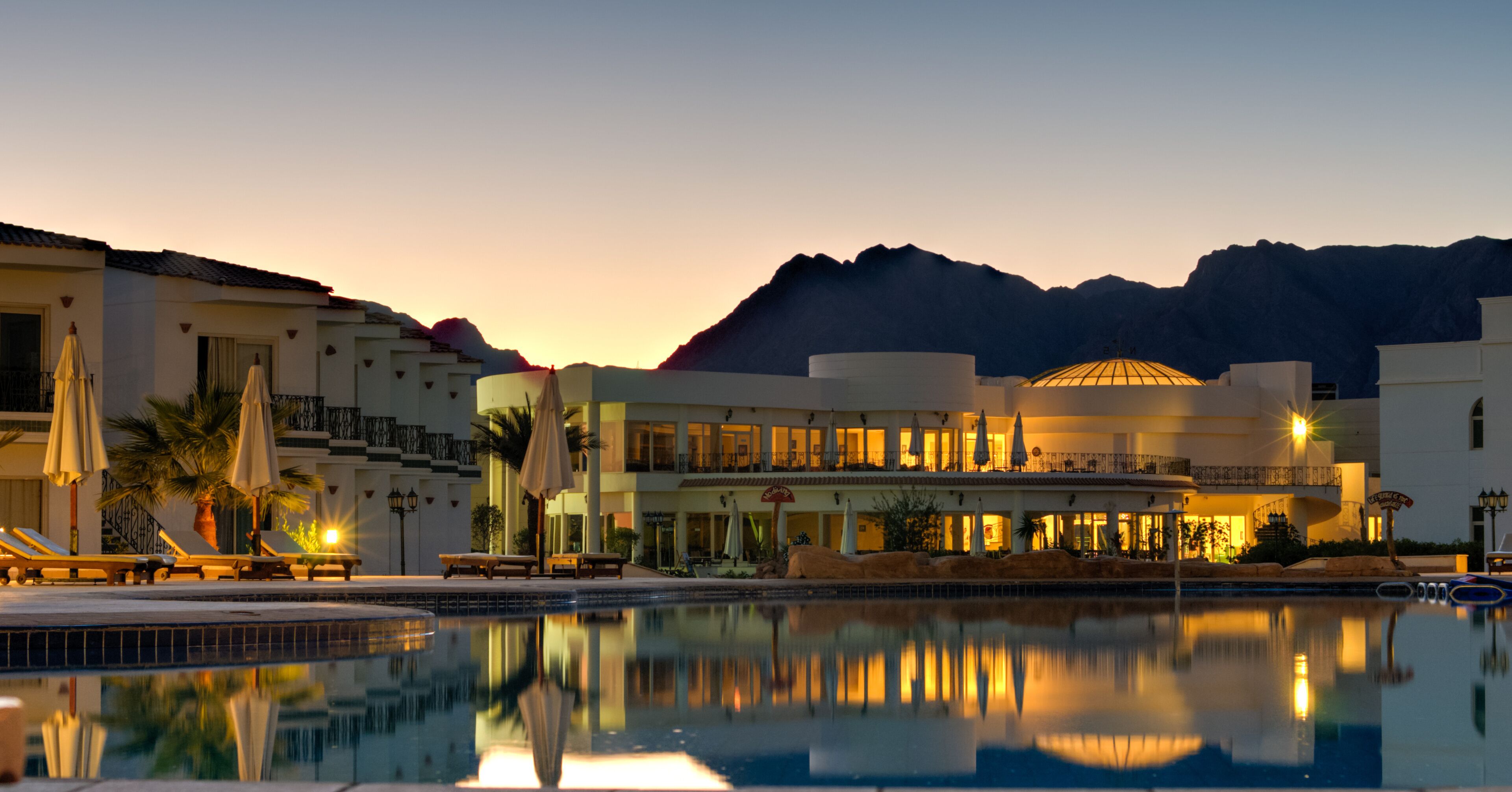 A resort with a swimming pool, lounge chairs, umbrellas, and a mountain backdrop at dusk.