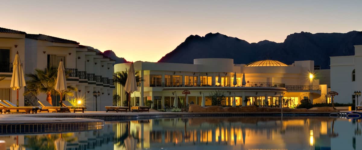 A resort with a swimming pool, lounge chairs, umbrellas, and a mountain backdrop at dusk.