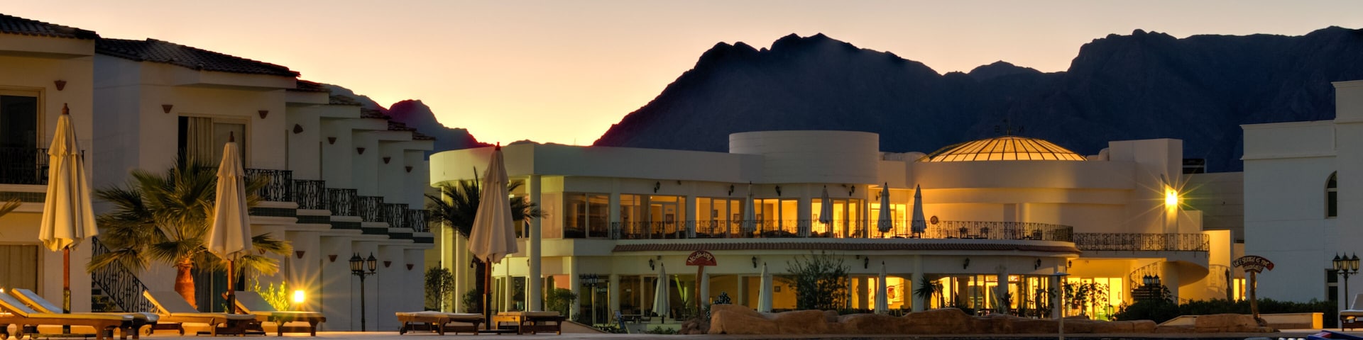A resort with a swimming pool, lounge chairs, umbrellas, and a mountain backdrop at dusk.