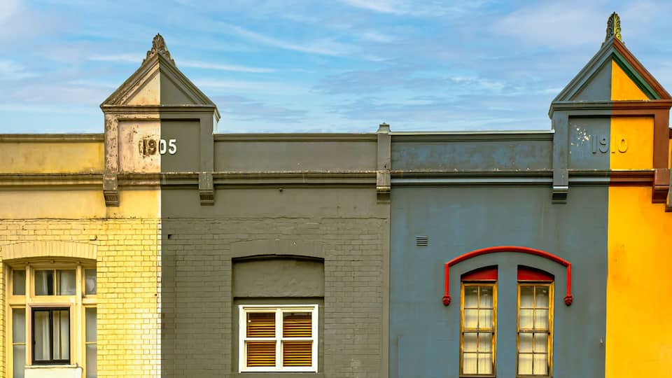 A row of buildings with distinct architectural styles, including a yellow facade with a red window trim and a grey building with a white window.