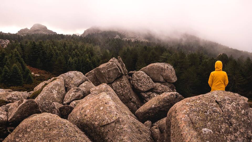 A person in a yellow jacket standing on large rocks overlooking a forested mountain range with fog.