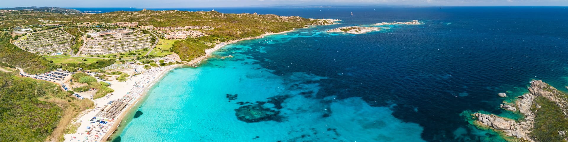 A coastal resort with a turquoise beach, clear waters, and a hilly landscape.
