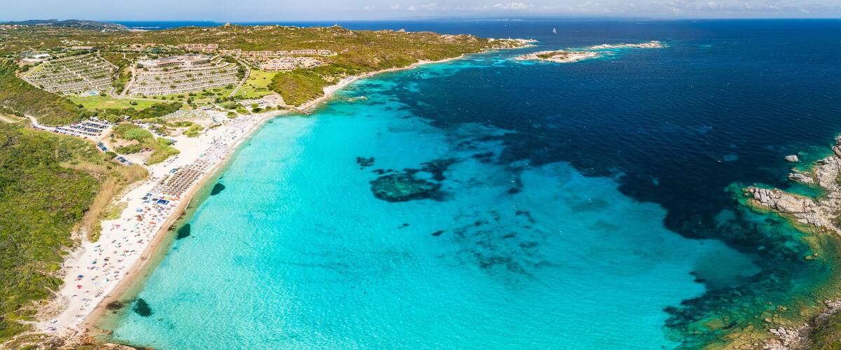 A coastal resort with a turquoise beach, clear waters, and a hilly landscape.