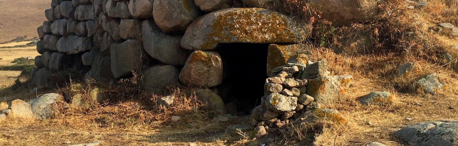 Una construcción de piedra con una entrada circular, rodeada de hierba seca y rocas.