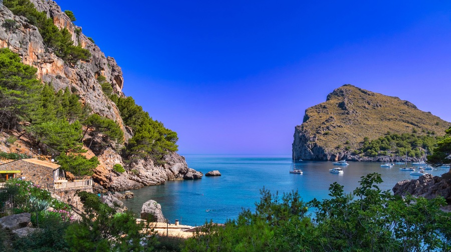 Un paisaje costero con acantilados rocosos, un mar azul cristalino y varias embarcaciones fondeadas cerca de la orilla.