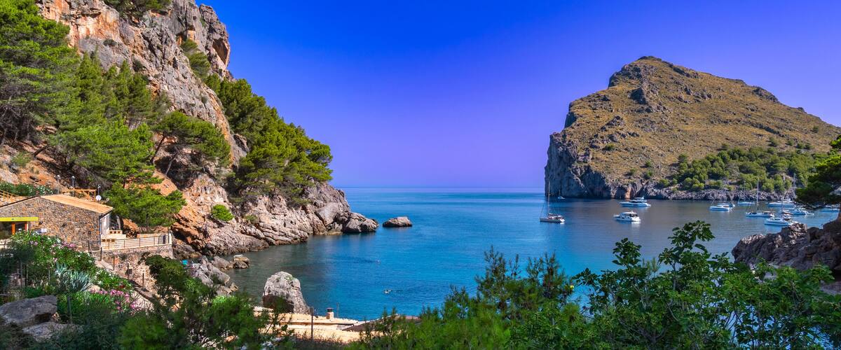A coastal landscape with rocky cliffs, a clear blue sea, and several boats anchored near the shore.