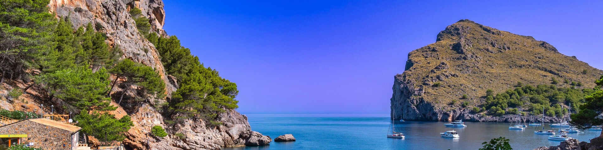 A coastal landscape with rocky cliffs, a clear blue sea, and several boats anchored near the shore.