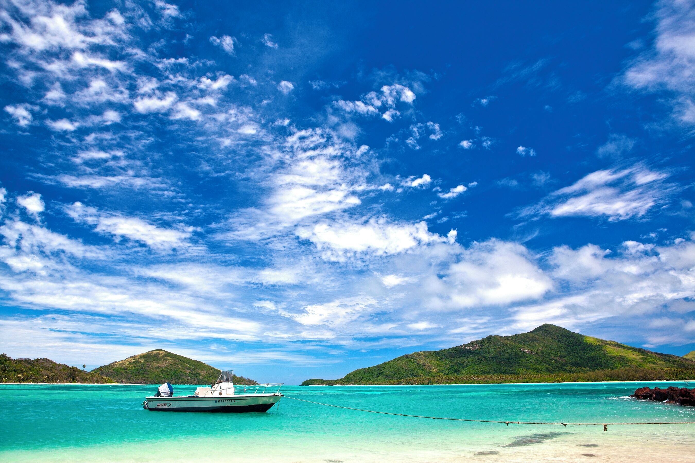 A boat anchored near a turquoise beach with clear skies and green hills in the background.
