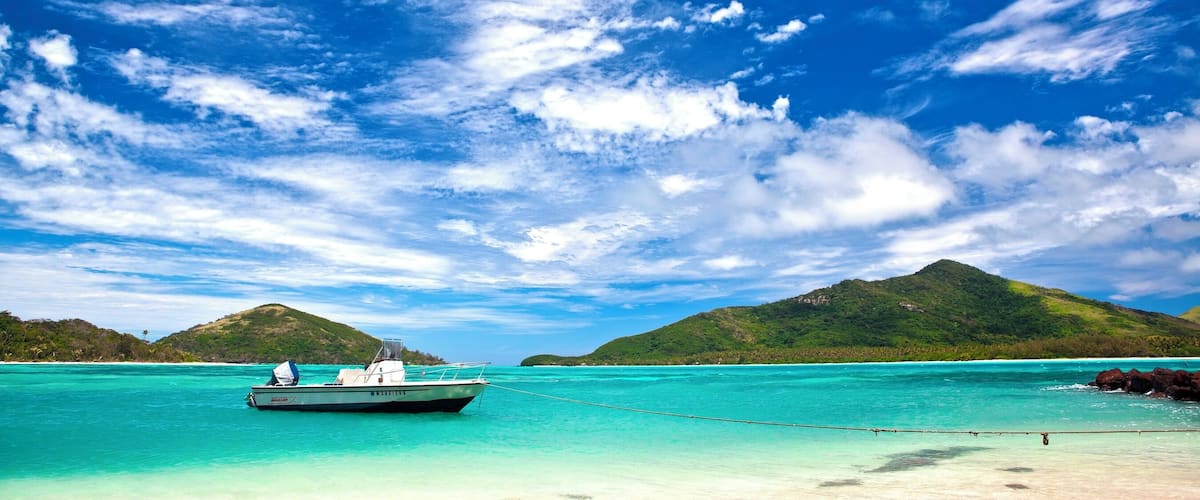 A boat anchored near a turquoise beach with clear skies and green hills in the background.