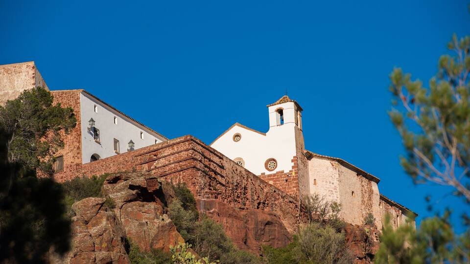 A white church with a bell tower, set against a clear blue sky, surrounded by rocky terrain and sparse vegetation.