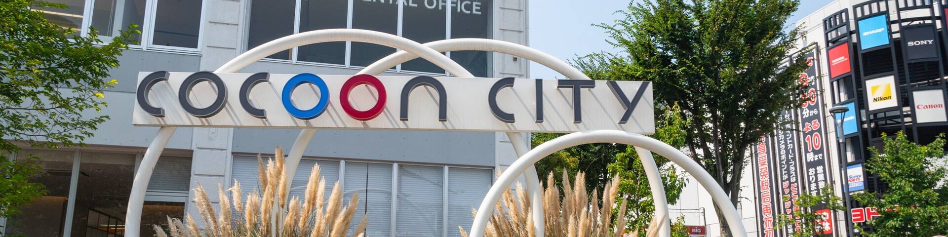 A sign for 'Cocoon City' with a circular design, surrounded by plants and a building in the background.