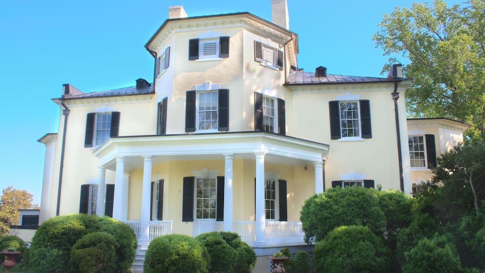 A large, two-story house with a wrap-around porch, multiple chimneys, and a well-maintained garden with trimmed bushes.