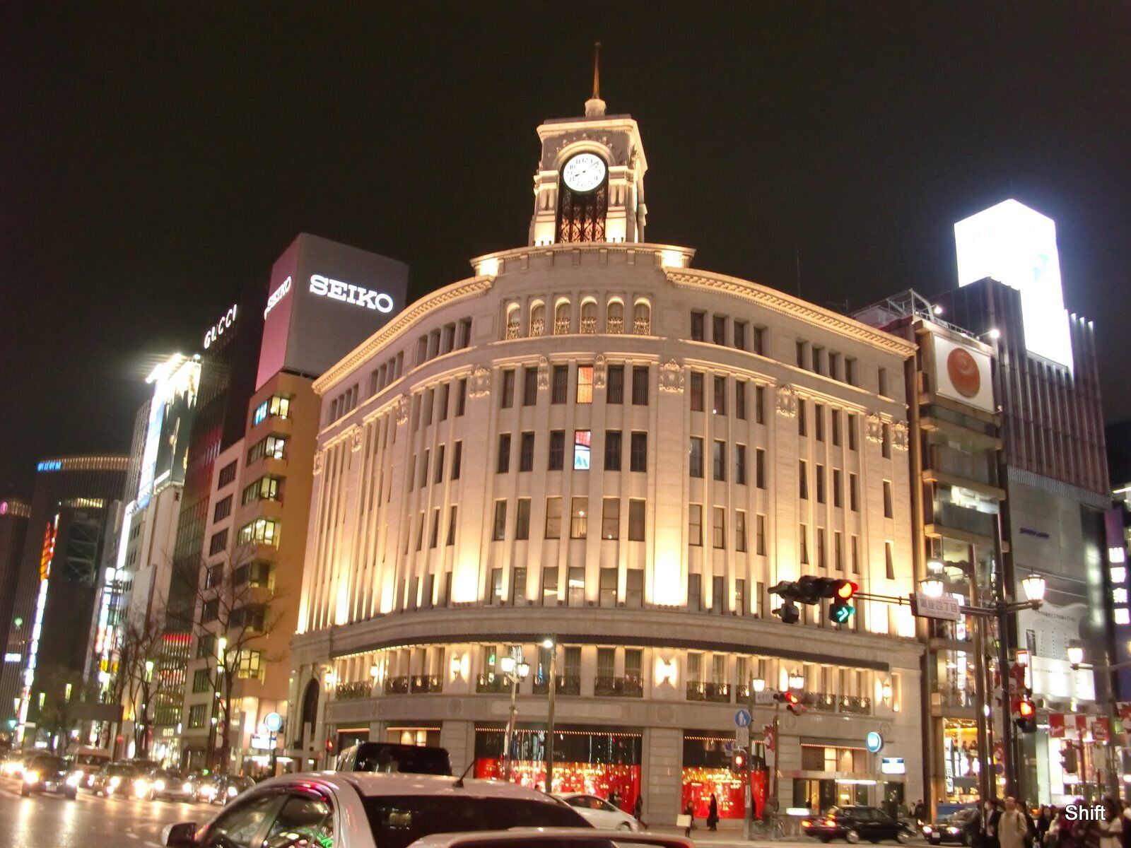 Um edifício histórico bem iluminado com uma torre do relógio, rodeado por arranha-céus modernos e tráfego intenso na rua.