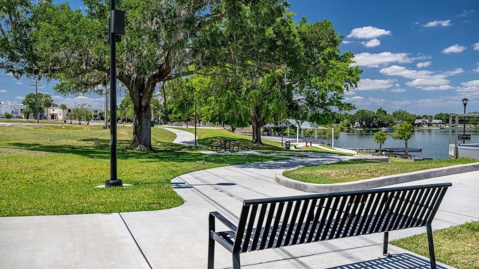 A park with a curved pathway, a bench, trees, a body of water, and a distant building.