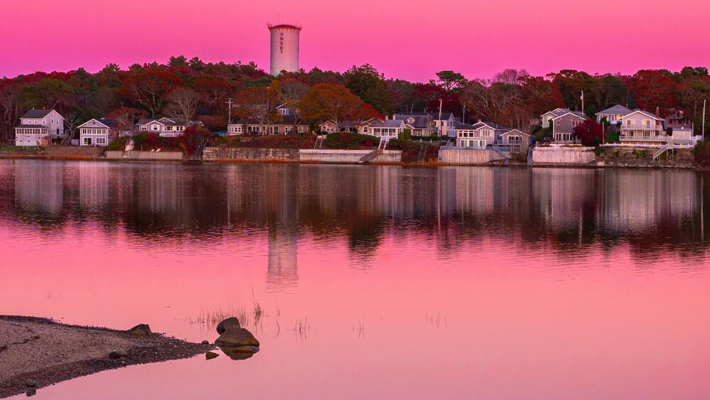 Un lac paisible, un ciel rose, une tour à eau et des maisons qui bordent la rive.