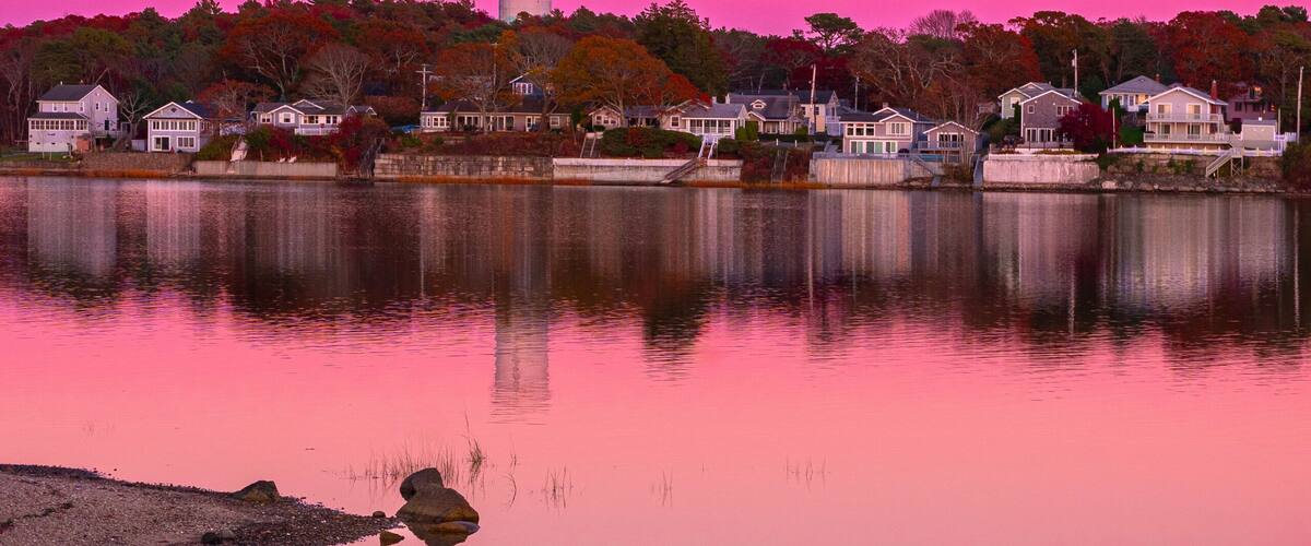A calm lake with a pink sky, a water tower, and houses along the shore.