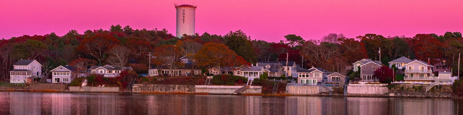 A calm lake with a pink sky, a water tower, and houses along the shore.