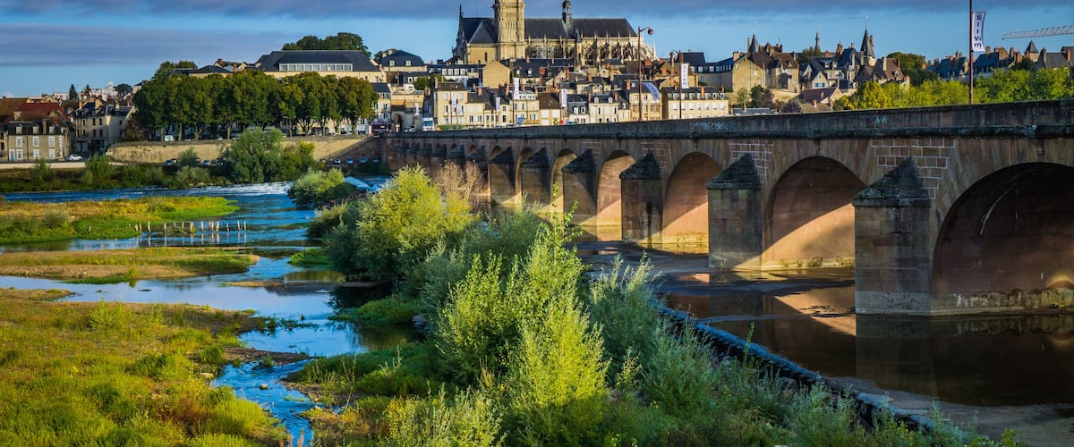 A stone bridge over a river, a historic church with a tower, and a town with buildings in the background.