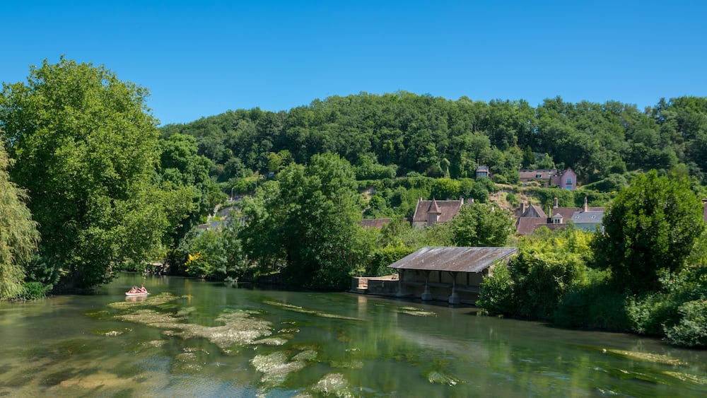Une rivière où flotte un petit bateau, entourée d’une végétation luxuriante et bordée de maisons sur le versant d’une colline.