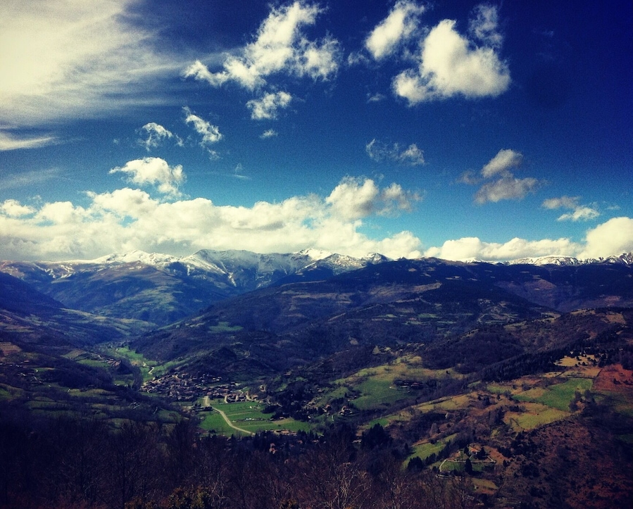 A mountainous landscape with snow-capped peaks, a valley with scattered houses, and a clear blue sky with clouds.