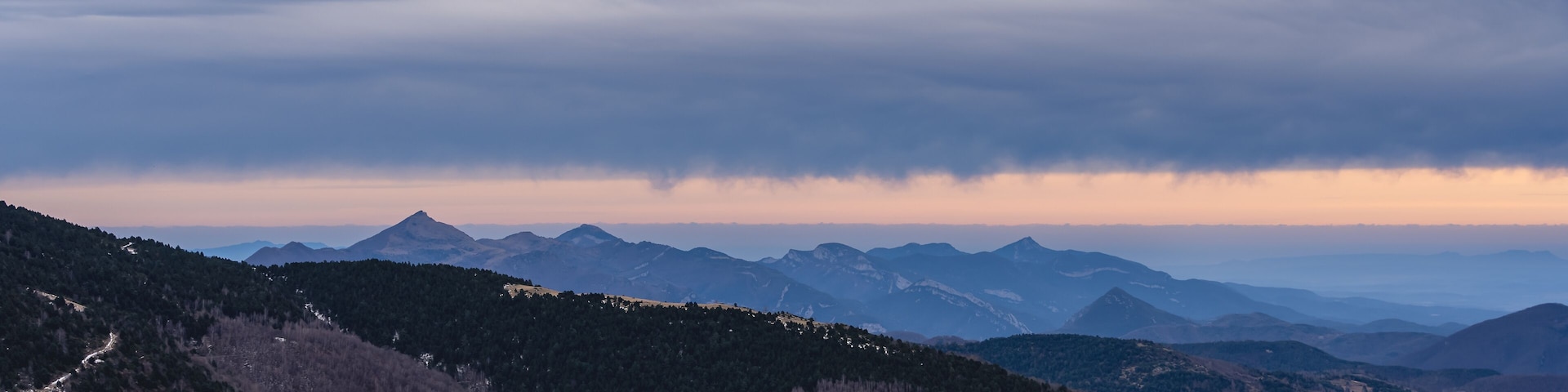 A mountainous landscape with a winding path and distant peaks under a cloudy sky.