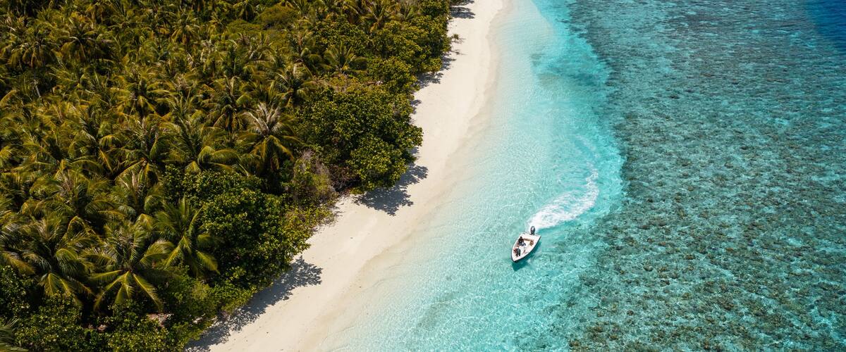 An aerial view of a beach with clear turquoise waters, a sandy shore, and dense tropical vegetation.