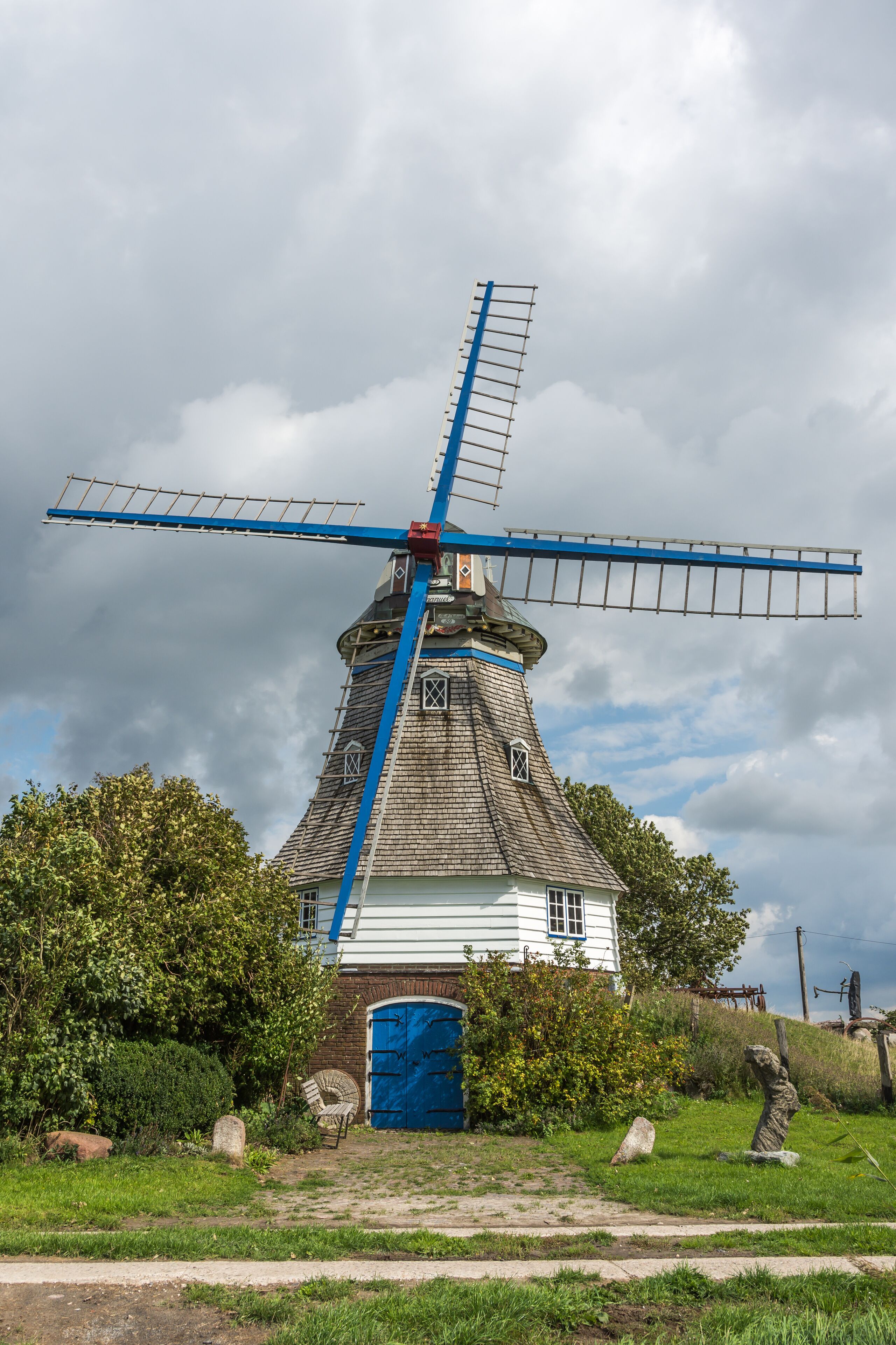 A traditional windmill with a blue and white structure, surrounded by greenery and a cloudy sky.