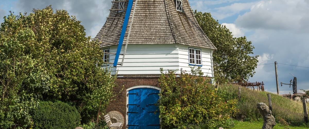 A traditional windmill with a blue and white structure, surrounded by greenery and a cloudy sky.