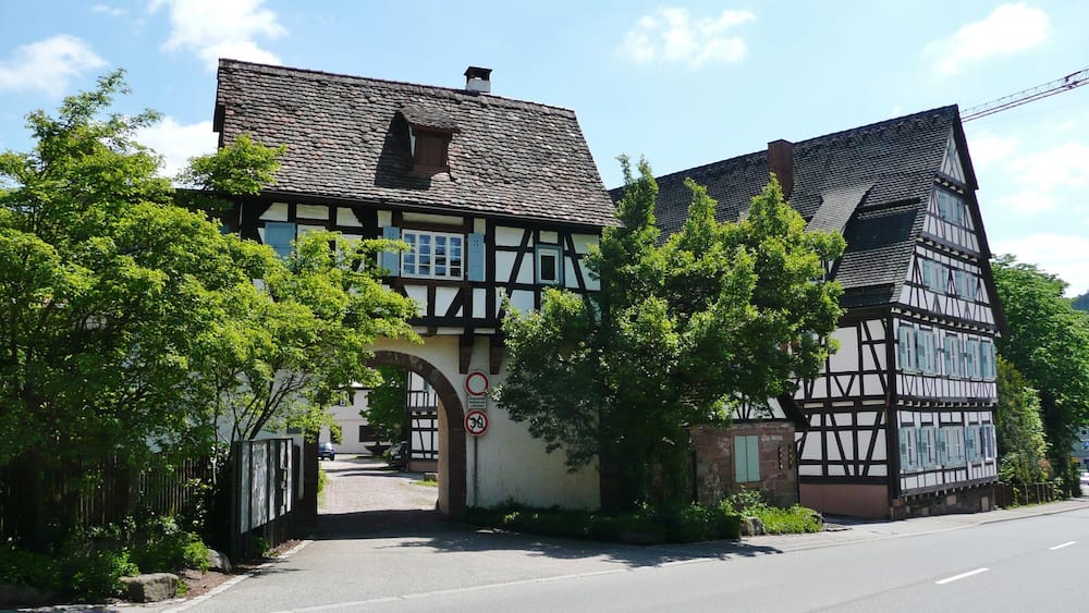 Une maison traditionnelle à colombages, surmontée d’un toit en pierre, entourée d’une végétation luxuriante et baignée d’un ciel dégagé.