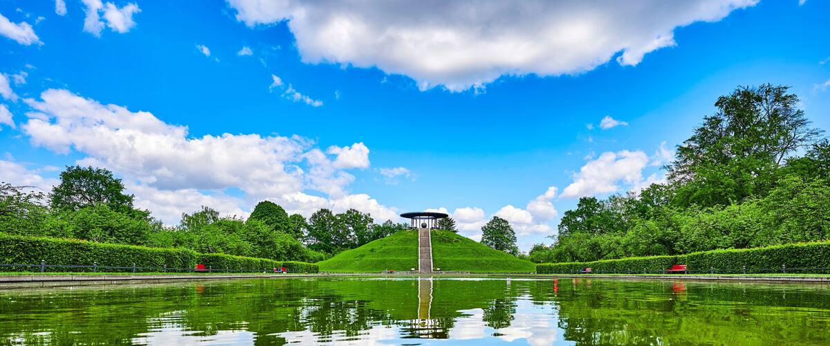 A park with a central monument, a pond reflecting the sky, and lush green trees.