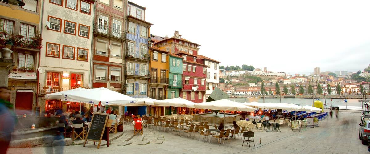 A bustling riverside square with colorful buildings, outdoor cafes, and a view of the cityscape in the background.