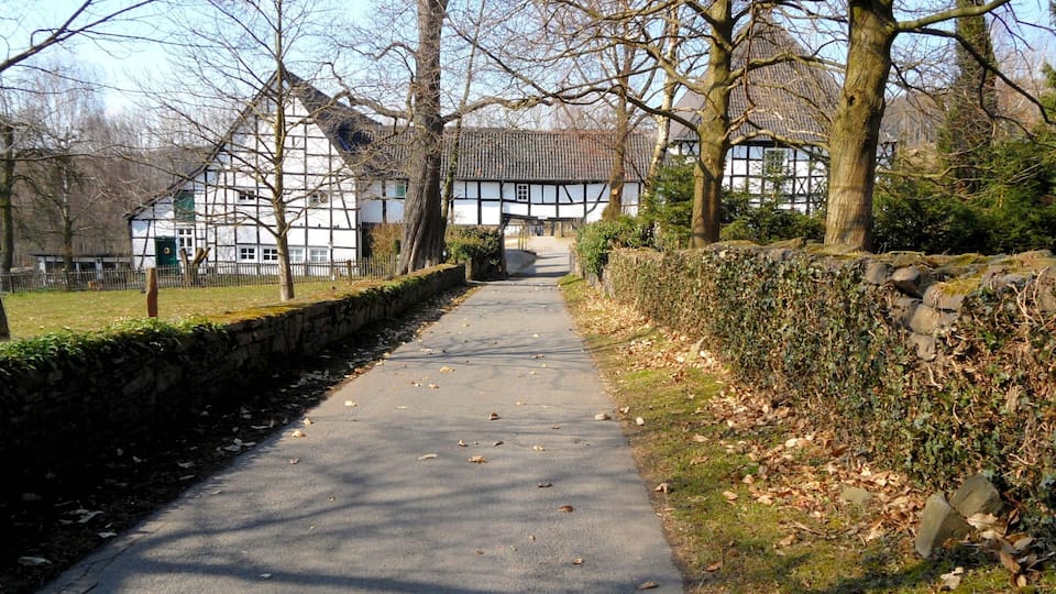 A pathway leading to a traditional half-timbered house with a thatched roof, surrounded by trees and a stone wall.
