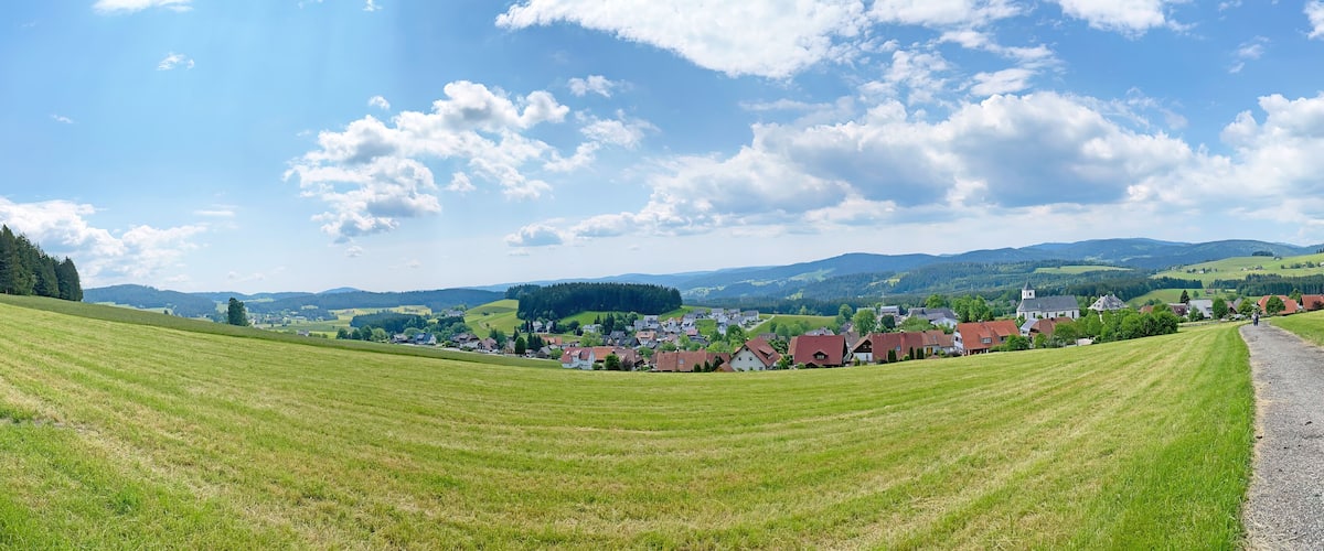 A rural landscape with rolling green fields, a village with red-roofed houses, and distant hills under a partly cloudy sky.
