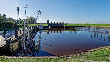 A calm river with a lock system, green grass, and wind turbines in the background.