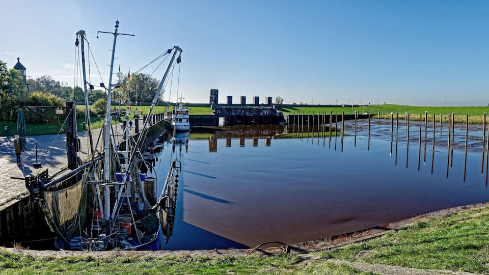 A calm river with a lock system, green grass, and wind turbines in the background.