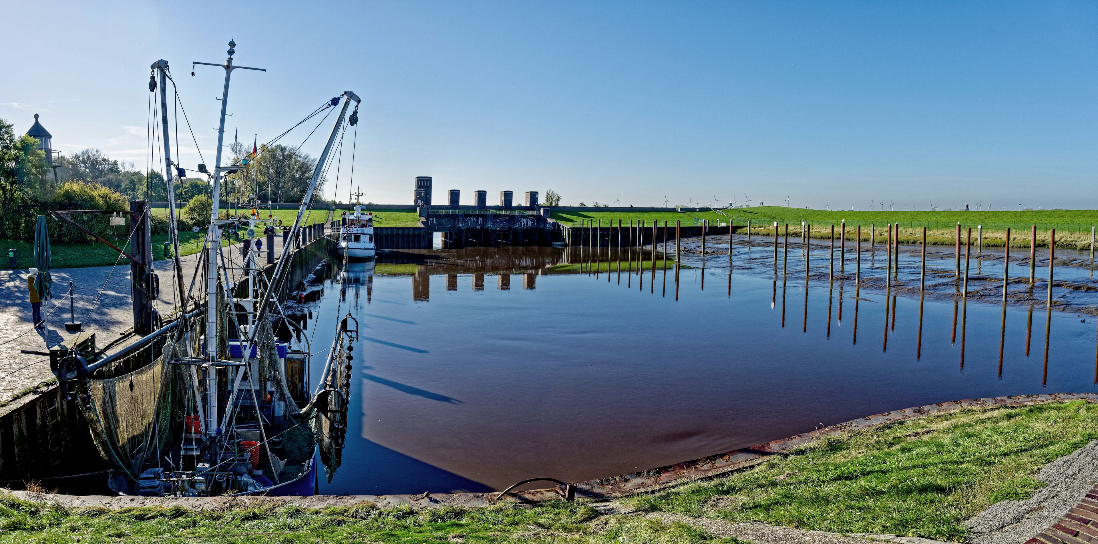 A calm river with a lock system, green grass, and wind turbines in the background.