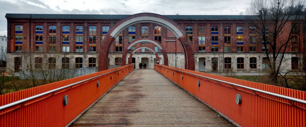A wooden bridge with orange railings leading to a large brick building with multiple windows and arches.