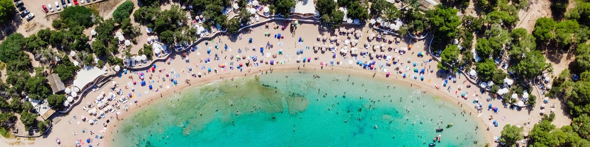 An aerial view of a beach with clear turquoise waters, a rocky shoreline, and a line of umbrellas and beach chairs along the edge.