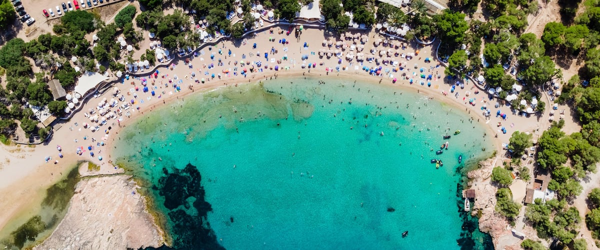 Vista aérea de una playa con aguas turquesas cristalinas, un litoral rocoso y una hilera de sombrillas y sillas de playa a lo largo de la orilla.