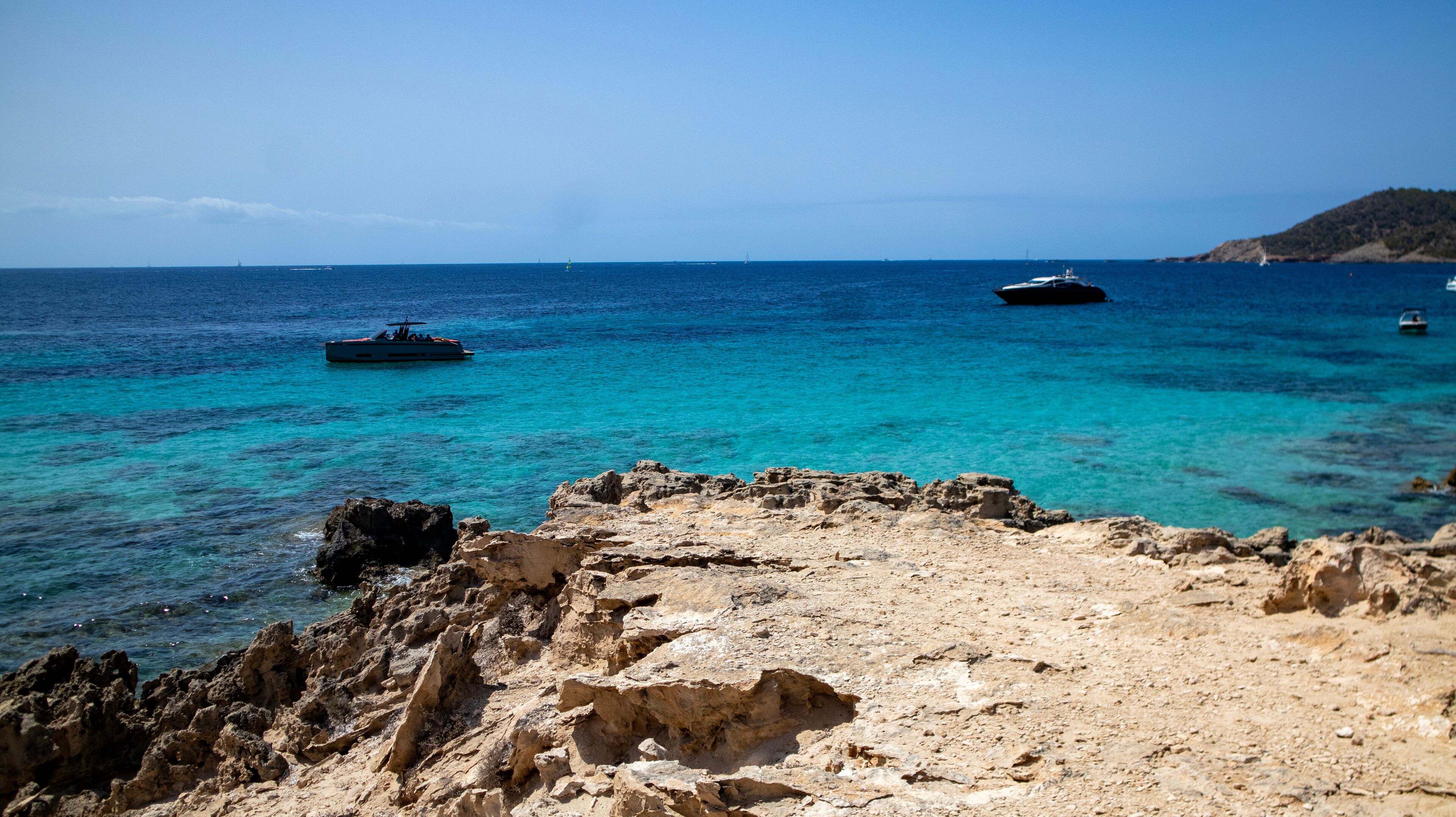 A rocky coastline with clear turquoise waters, boats, and a distant hill.